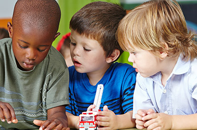 Three children in kindergarten playing with building blocks and cars